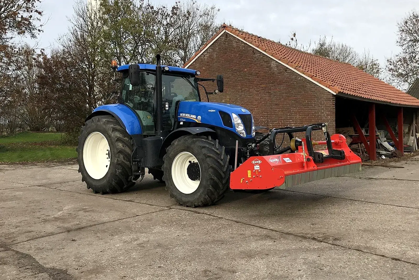 New Holland trekker met rode Kuhn klepelbak voor een schuur op het erf
