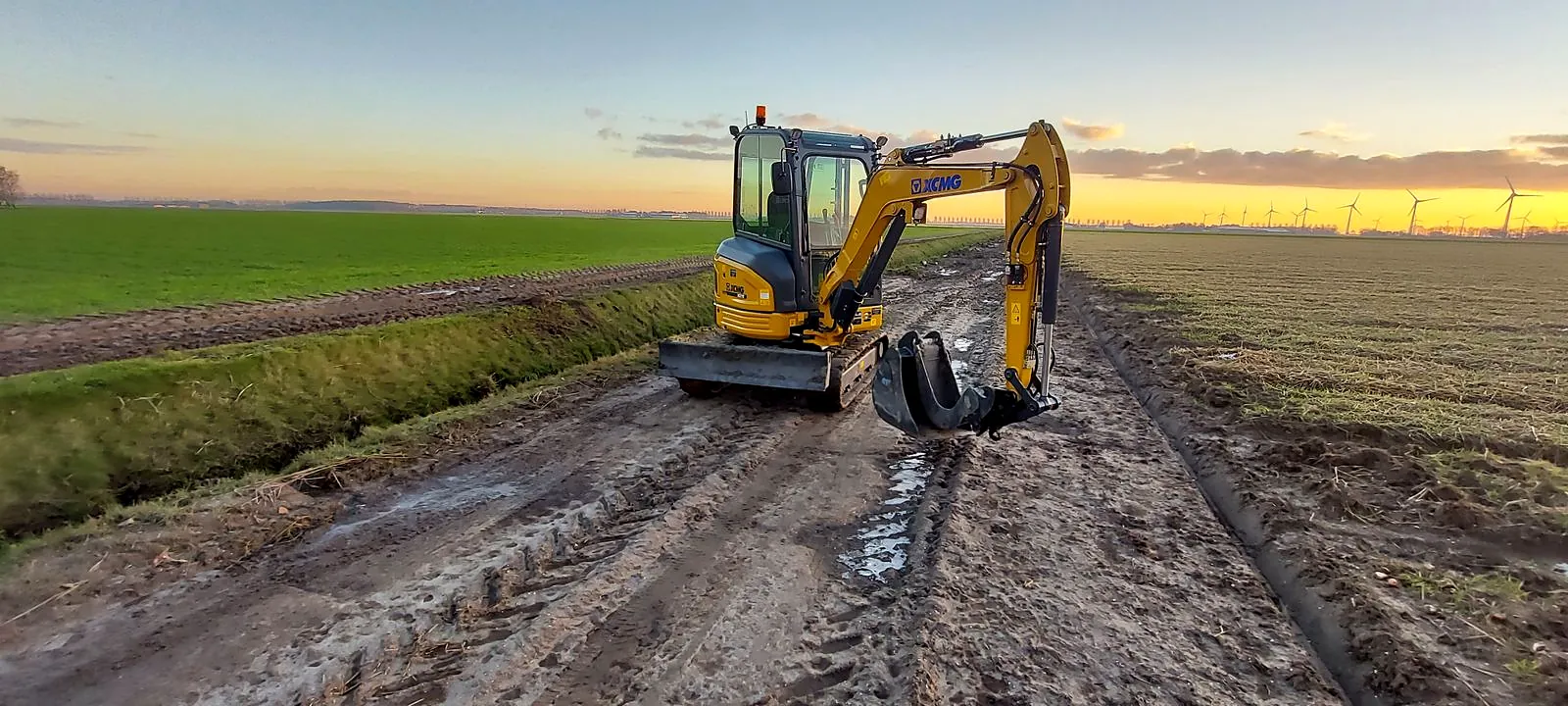 XCMG minikraan op een nat werkpad langs een sloot bij zonsondergang